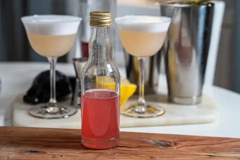 En flaska med rosa rabarbersirap står framför två drinkar med skum. En shaker och en citronklyfta syns i bakgrunden.
