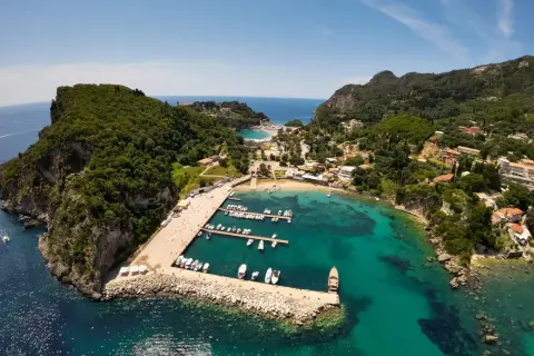 Malownicza zatoka na Korfu, idealna na wakacje z dzieckiem. Widać tu port z łodziami, plażę i zielone wzgórza.