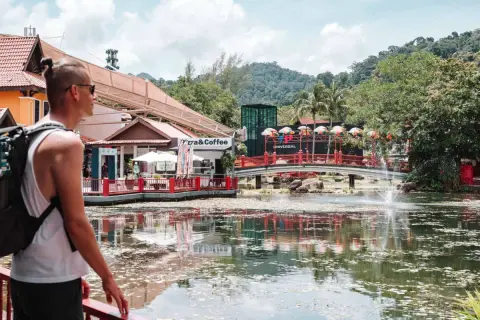 Mann mit Rucksack, der die bunte Malaysia Kleidung und die tropische Landschaft genießt. Ein See mit Fontäne und Brücke.