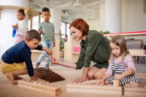 Niños y maestra en el suelo, aprendiendo con materiales didácticos. Las rutinas diarias en el aula fomentan el desarrollo.
