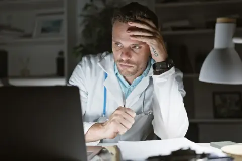 Lekarz z rezygnacją opiera głowę na dłoni, pracując po godzinach. Widać zmęczenie i frustrację.