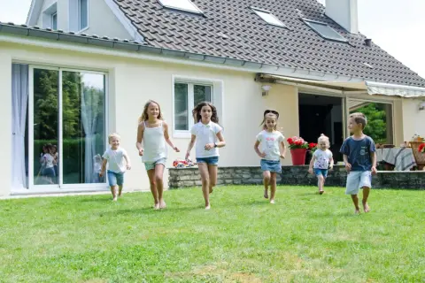 Dzieci biegają po trawniku przed domem z jasnym tynkiem zewnętrznym. Różne rodzaje tynków zewnętrznych mogą nadać budynkowi unikalny wygląd.