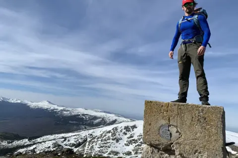 Hombre con mochila en la cima de un pico nevero, con vistas a montañas nevadas bajo un cielo azul con nubes.