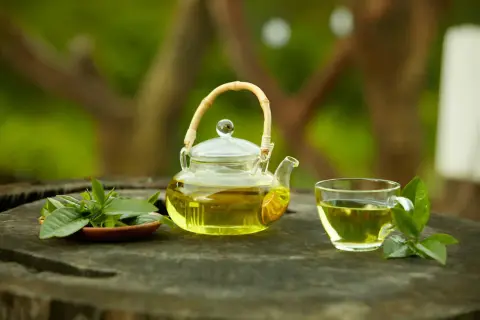 Une théière en verre remplie de thé vert, une tasse assortie et des feuilles de thé frais sur une table en bois.