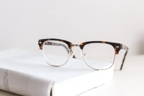 Okulary z oprawkami w stylu retro, idealne do korygowania wady wzroku +2. Leżą na białej książce.