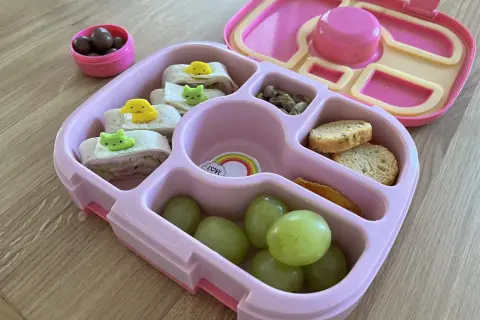 Różowa śniadaniówka do szkoły z przegródkami zawiera zawinięte tortille z uroczymi ozdobami, winogrona, orzeszki pistacjowe, krakersy i suszone mango.