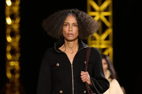 Una mujer con cabello rizado y gris camina por una pasarela, luciendo un elegante cárdigan negro con botones dorados, como una **chanel cantante**.