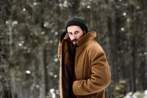 Mężczyzna w beżowym płaszczu i czarnej czapce, prezentujący stylowe zimowe stylizacje męskie na tle zaśnieżonego lasu.