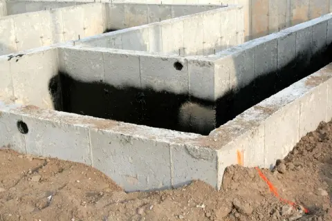 Betonowe bloczki zalewowe tworzą fundament domu, zabezpieczone czarną izolacją przeciwwilgociową.
