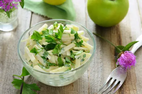 Frischer Kohlrabi Salat mit Petersilie in einer Glasschüssel, garniert mit einer lila Blume und grünen Äpfeln im Hintergrund.