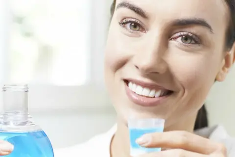 Woman smiling, holding a bottle of blue mouthwash and a cap filled with mouthwash.