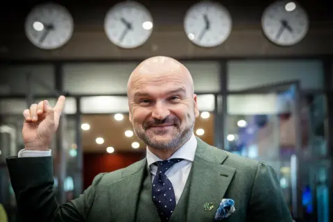 Rafał Brzoska, z uśmiechem i uniesionym palcem, symbolizuje swój rosnący majątek. Za nim zegary pokazują różne strefy czasowe.