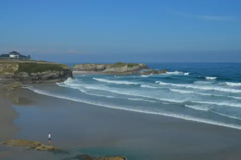 La **playa Os Castros** se extiende bajo un cielo azul, con olas rompiendo en la arena dorada. Una casa moderna corona el acantilado.
