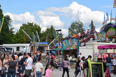 Buntes Treiben auf einem Volksfest mit Fahrgeschäften und vielen Besuchern. Ein Schiff-Karussell dreht sich, während Menschen an Ständen vorbeischlendern.