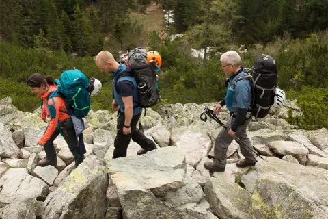 Drei Wanderer mit Rucksäcken und Helmen auf felsigem Untergrund. Die **Packliste Klettersteig** ist gut gefüllt für das Abenteuer.