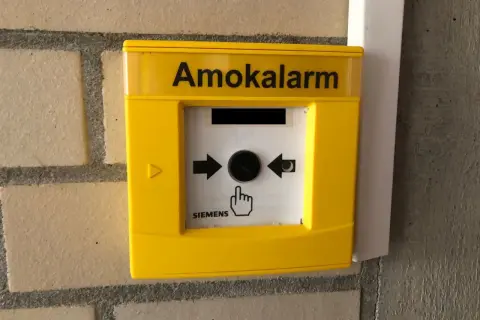 Gelber Amokalarm-Knopf an einer Ziegelwand. Ein Symbol zeigt eine Hand, die auf den Knopf drückt, um einen Amoklauf an einer Schule zu melden.