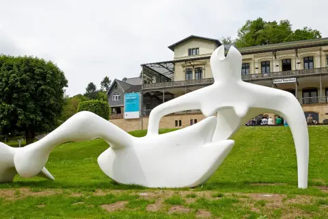 Weiße Skulptur einer liegenden Figur im Freien vor einem Gebäude mit der Aufschrift "Bahnhof Rolandseck".