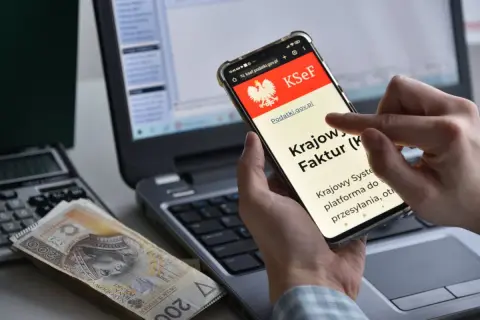 Dłoń trzyma smartfon z ekranem KSeF, obok leżą pieniądze i kalkulator.