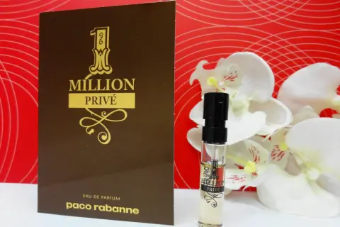 Złoty flakonik i próbka Paco Rabanne 1 Million Privé na tle czerwonych fal i białych kwiatów.