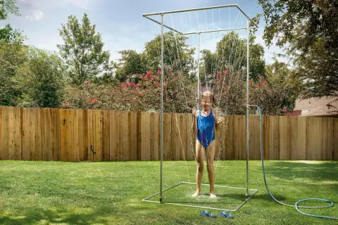 Ein Mädchen im blauen Badeanzug genießt eine erfrischende Dusche unter einer selbstgebauten Gartendusche im sonnigen Garten.