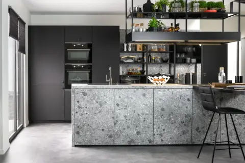 Cuisine moderne aux façades grises et à l'îlot central en sol terrazzo. Électroménagers intégrés et étagères noires remplies d'objets décoratifs.