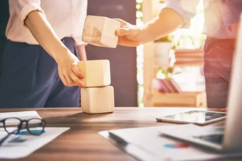 Dwie osoby wspólnie budują wieżę z drewnianych klocków, symbolizując budowanie zespołu.