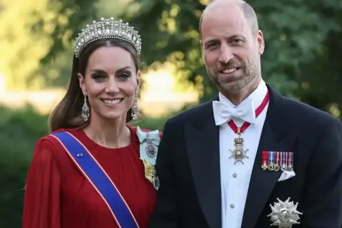 Kate i William w królewskich strojach, z diademem i medalami, uśmiechają się do zdjęcia.