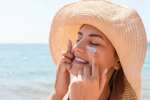 Une femme souriante applique de la crème solaire sur son visage, protégeant sa peau. Est-ce que la crème solaire hydrate ? Oui, certaines le font !