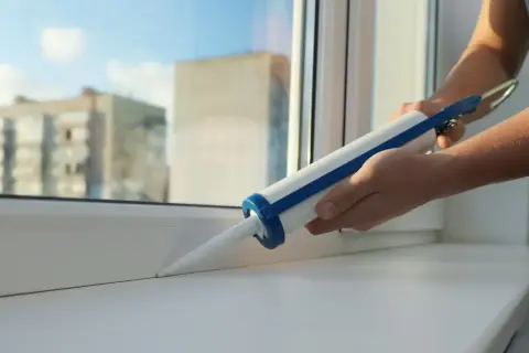 Ręce trzymają pistolet z uszczelniaczem, aplikując go na biały parapet okna.