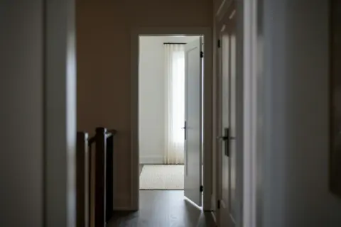 Korytarz z otwartymi drzwiami prowadzącymi do jasnego pokoju z zasłonami. Widoczny fragment balustrady schodów. 193 kk.