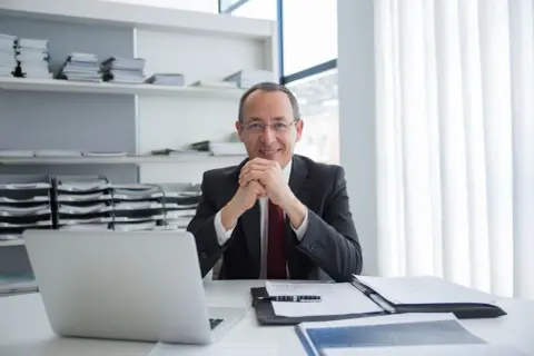 Uśmiechnięty mężczyzna w garniturze siedzi przy biurku z laptopem i dokumentami. Jego mała działalność gospodarcza prosperuje.