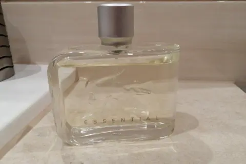 Szklana butelka perfum Lacoste Essential Sport z srebrną nakrętką, stojąca na blacie.