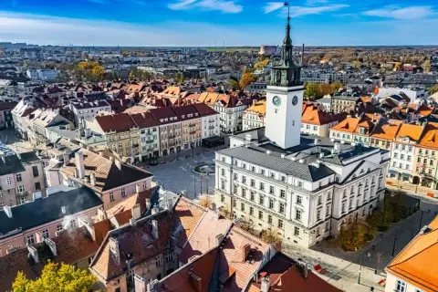 Widok z lotu ptaka na rynek z ratuszem i wieżą zegarową, otoczony kamienicami.