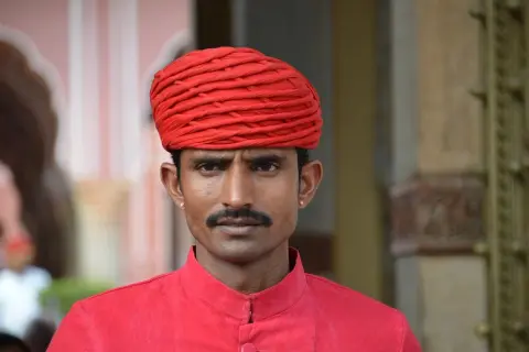Un homme porte un turban rouge vif et une chemise assortie. Sa moustache fine souligne son regard intense.