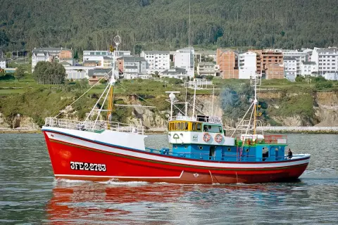 El barco museo boniteiro Reina del Carmen navega con su casco rojo y azul, reflejándose en el agua.