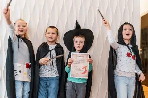 Cztery dzieci w strojach czarodziejów z różdżkami. Jeden chłopiec w kapeluszu czarownicy trzyma dyplom.
