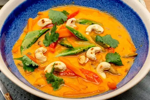Ein cremiges rotes Thai Curry mit Cashewkernen, Zuckerschoten und Karotten in einer blauen Schüssel.