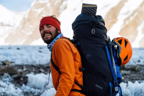 Kacper Tekieli, mit roter Mütze und orangefarbener Jacke, lächelt in die Kamera, bereit für sein nächstes Bergabenteuer.
