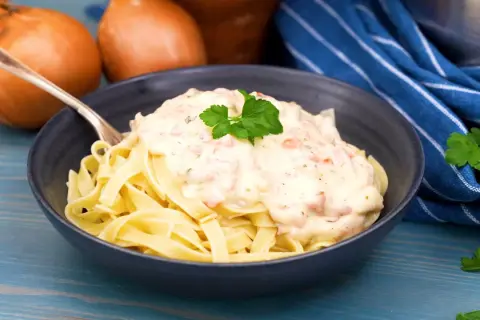 En skål med tagliatelle toppad med krämig ost och skinksås, garnerad med persilja. Två gula lökar syns i bakgrunden.
