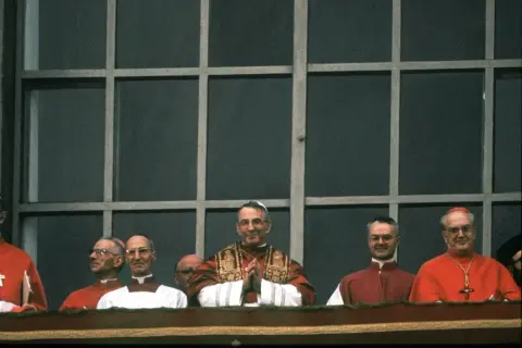 Uroczyste zgromadzenie duchownych, wśród nich papież Jan Paweł II. Wszyscy w strojach liturgicznych, przypominają o ważnych imionach papieży.
