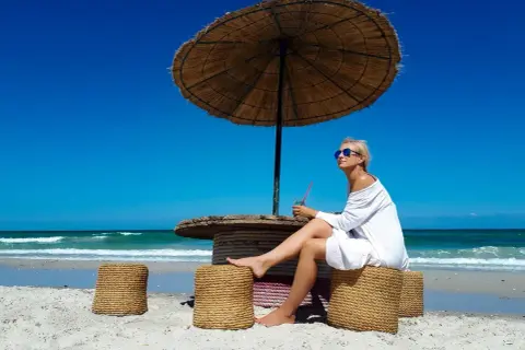 Kobieta w białej sukience siedzi pod parasolem na plaży na Djerbie. Uważać na słońce i upał.