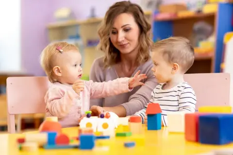 Dzieci bawią się klockami przy stole, a opiekunka patrzy z uśmiechem. Dofinansowanie do żłobka ułatwia rodzicom opiekę nad maluchami.
