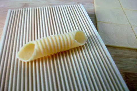 Frische Pasta selbst machen ohne Maschine: Ein geriffeltes Werkzeug formt Teig zu Röhren. Daneben liegen Teigplatten.