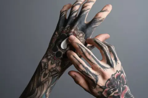 Hände mit detaillierten Tattoos werden eingecremt. Eine Person trägt eine weiße Creme auf ihre tätowierten Finger und Handrücken auf.