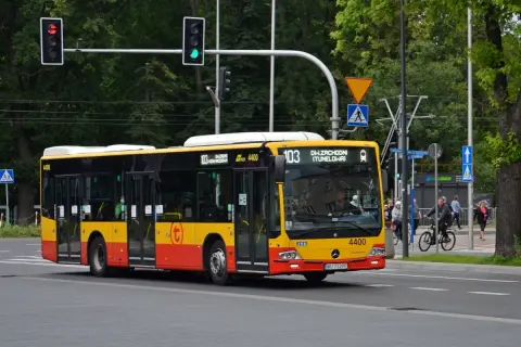 Żółto-czerwony autobus linii 103 w Warszawie jedzie ulicą. W tle drzewa i sygnalizacja świetlna.