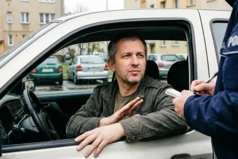 Policjant wypisuje mandat karny kierowcy, który siedzi w samochodzie i gestykuluje.
