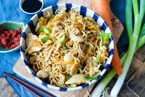 Bami Goreng mit Hühnchen, Karotten und Frühlingszwiebeln in einer Schüssel, daneben Sojasauce, Chili und frisches Gemüse.