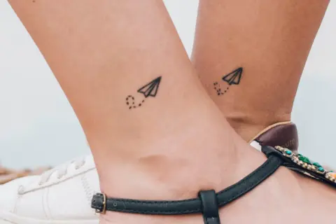 Dwa małe tatuaże samolotów z kropkowanym śladem na kostkach. Symbolizują podróże i wolność.