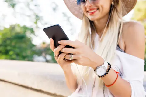 Uśmiechnięta kobieta w kapeluszu słucha muzyki przez słuchawki, trzymając telefon. Może szuka instrukcji, jak zainstalować radio w telefonie.