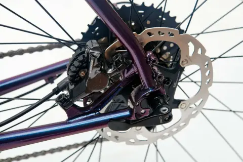 Błyszcząca, fioletowa rama roweru z widocznym hamulcem tarczowym Shimano i kasetą. Sztywna oś w rowerze zapewnia stabilność.